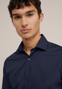 Temno modra srajca z gumbom na ovratniku, iz gladkega materiala, prilegajoče zasnove in diskretno gumbnico na sprednji strani.