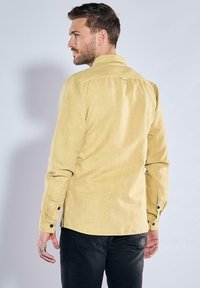 Helles gelbes Langarmshirt mit Knopfmanschetten, einem Rückenpasse und einem leicht strukturierten Stoff. Tragen mit dunklen Hosen.