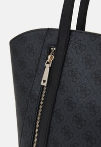 Gros plan sur un sac fourre-tout noir avec un motif monogramme, fermeture éclair dorée détaillée et doubles poignées noires.