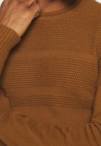 Brązowy sweter wełniany z różnorodnymi teksturowymi wzorami, z prążkowanym okrągłym dekoltem i długimi rękawami, podkreślający przytulny i ciepły wygląd.