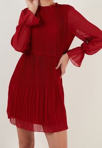 Abito rosso a pieghe con maniche lunghe e trasparenti e un colletto a ruches. Presenta una vita elastica e un tessuto fluido che si apre delicatamente all'orlo.