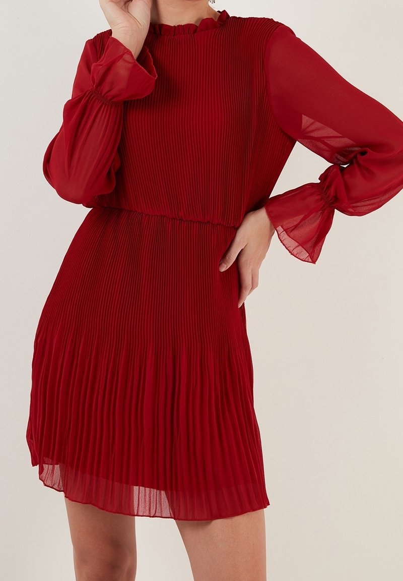 Abito rosso a pieghe con maniche lunghe e trasparenti e un colletto a ruches. Presenta una vita elastica e un tessuto fluido che si apre delicatamente all'orlo.