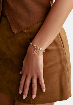 Silberner Gliederarmband mit einem pinken Bärenanhänger und einer runden Metallplakette, getragen an einer Hand vor einem braunen Wildlederrock.