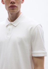 Fehér póló sima pamutból, kötött gallérral, két gombos plackettel és a mellkason hímzett "BOSS" logóval.