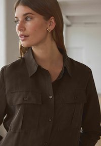 Brown button-up shirt made of a smooth fabric, featuring a collar and two chest pockets with button closures. Notable simplicity in design.