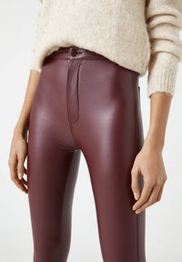 Kvinna som bär högmidjade, vinröda läderbyxor med knappstängning och en beige fluffig tröja, med händerna avslappnat längs sidorna.