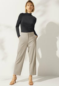 Frau in schwarzem Langarm-Turtleneck, beigen hoch geschnittenen, weiten Hose und tanfarbigen, geschnittenen High-Heels, die mit den Händen in den Taschen steht.