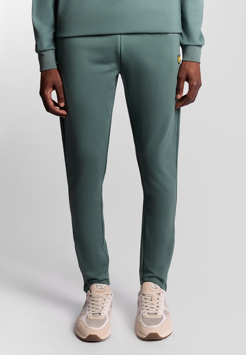 Groene sportieve joggingbroeken van gladde stof, met zijnaad, een aansluitende pasvorm en een klein logopatch nabij de heup.