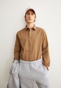 Camicia marrone con bottoni e un logo discreto, abbinata a pantaloni della tuta grigi. Il cappello è rosa chiaro, con un logo sulla parte frontale.