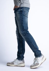 Dopasowane niebieskie dżinsy z wyblakłym praniem, pięć kieszeni i szczegóły przeszycia, w połączeniu z białymi i szarymi sneakersami w stylu retro.