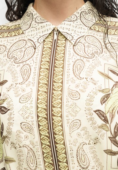 Primer plano de una camisa beige con patrones marrones de cachemira y flores, que presenta un cuello puntiagudo y una cremallera central.