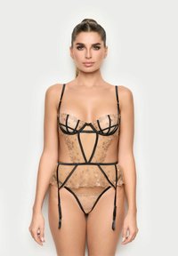 Mujer vistiendo lencería beige transparente con tirantes negros y bordados de estrellas, de pie frente a un fondo claro, mirando hacia adelante.