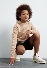 Felpa beige con tasca a marsupio e logo bianco, pantaloni sportivi neri in rete, e sneaker bianche con dettagli rossi e motivo decorativo.
