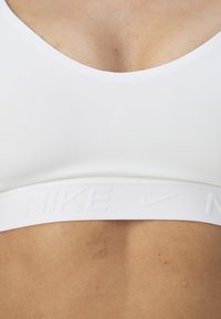 Hvit sports-bh med en jevn tekstur, med en bred band preget med Nike-logoen. V-halsdesign gir en tettsittende passform.
