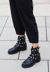 Stivaletti neri in pelle con decorazioni di perle e cinturini con fibbia, indossati con pantaloni neri a vita alta su un pavimento piastrellato.