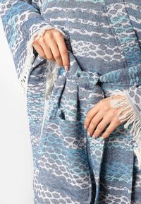 Modra in bela obleka z vzorcem s frufrujem na rokavih, ki vsebuje abstraktno zasnovo in kravato v pasu, izdelana iz teksturirane tkanine.