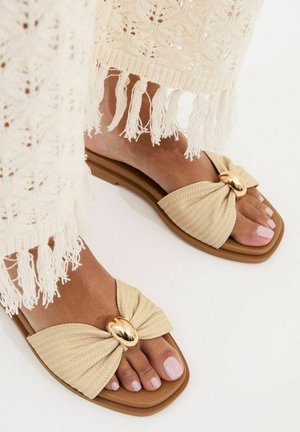 Pies con sandalias tipo slide beige con detalle de hebilla dorada, vistiendo pantalones de crochet color crema con flecos en el dobladillo.