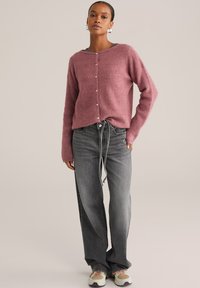 Femme portant un pull boutonnière rose, un jean noir délavé ample avec une ceinture à nouer, des baskets blanches, se tenant devant un fond uni.