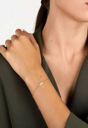 Wrist with delicate gold chain bracelet featuring three small white pearls, resting on shoulder over olive green V-neck garment.