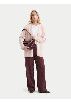 Femme portant un cardigan rose clair, une chemise blanche, un pantalon large bordeaux, des chaussures foncées, et tenant un sac à main en cuir marron foncé.