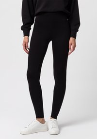 Černé legíny vyrobené z pružného materiálu, přiléhavý střih, vysoký pas, hladká textura, spárované s bílými teniskami a černým topem s dlouhým rukávem.