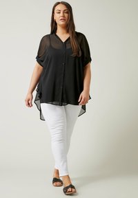 Kobieta idąca, ubrana w przezroczystą czarną luźną bluzkę z guziczkami na czarnym topie, białe dopasowane spodnie i czarne sandały na otwartym palcu.