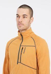 Oranger Fleece-Pullover mit hohem Kragen, halbem Reißverschluss und Kontrastnähten. Verfügt über eine Brusttasche mit Reißverschluss.