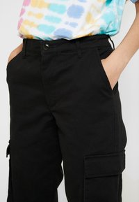 Černé cargo kalhoty s bočními kapsami a knoflíkovým zapínáním. Tkanina vypadá odolně s hladkým povrchem. Noseny s multikolorním tričkem s efektem tie-dye.