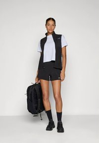 Zwart vest, lichtblauw cropped t-shirt, zwarte shorts en zwarte sneakers. Houdt een zwarte rugzak met meerdere vakken vast.