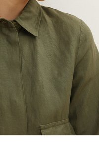 Camicia verde oliva con bottoni realizzata in tessuto testurizzato. Presenta un colletto e una tasca sul petto, con una vestibilità leggermente larga e cuciture visibili.