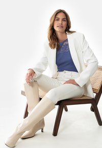 Blazer blanc sur un haut en dentelle bleu, associé à un jean blanc et des bottes en cuir beige à hauteur de genou. Modèle assis sur une chaise en bois.