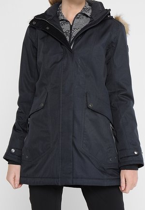 Femme portant une longue veste d'hiver noire avec des poches avant boutonnées et zippées, une capuche et des poignets boutonnés, par-dessus une chemise à motifs.