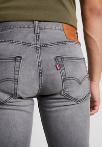 Grå denimjeans med slim fit, med två bakfickor, en läderlogotyp och en röd etikett på vänster ficka.