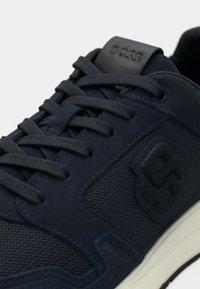 Σκούρο μπλε αθλητικό παπούτσι με υφασμάτινα και σουέντ υλικά, με επίπεδες κορδόνια, ανάγλυφο λογότυπο και λαστιχένια σόλα με αντίθετη κρέμ λεπτομέρεια.