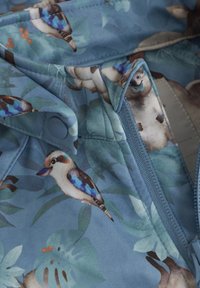 Blått tyg med ett naturmotiv av fåglar och blad, dragkedjestängning med en blå dragkedja och cirkulär knapppdetalj. Mjuk textur.