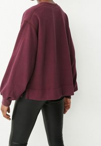 Sweat-shirt pull maroon à manches larges, avec des poignets côtelés et des fentes sur les côtés. Assorti à un pantalon noir ajusté. Texture lisse.