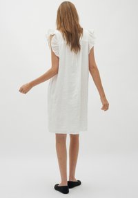Weißes Leinenkleid mit Rüschensleeves, lockerer Passform, knielangem Saum und strukturiertem Aussehen. Getragen mit schwarzen Ballerinas.