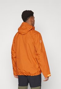Veste orange résistante à l'eau avec capuche, ourlet élastique et poignets. Présente un logo sur la manche et une texture lisse.