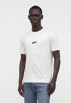 Uomo che indossa una maglietta bianca semplice a manica corta con un piccolo emblema dorato sul petto e jeans azzurri chiari, in piedi davanti a uno sfondo bianco.