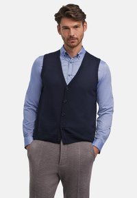 Gilet lavorato a maglia blu navy con scollo a V, sei bottoni sul davanti, indossato sopra una camicia azzurro chiaro con bottoni. Pantaloni grigi texture completano l'outfit.