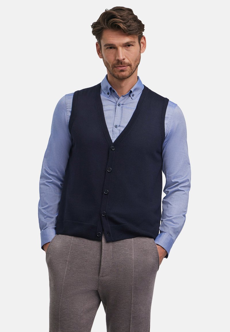 Gilet lavorato a maglia blu navy con scollo a V, sei bottoni sul davanti, indossato sopra una camicia azzurro chiaro con bottoni. Pantaloni grigi texture completano l'outfit.