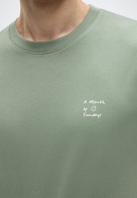 Heleroheline puuvillane T-särk ümmarguse kaelusega ja lühikeste varrukatega. Rinnal valge tikandiga tekst "A Month of Sundays".