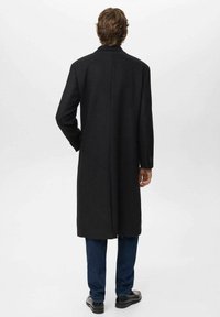 Hombre de pie de espaldas, con un abrigo largo negro, jeans azules oscuros y zapatos negros, sobre un fondo claro y uniforme.