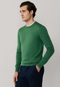 Grüner Pullover mit rundem Ausschnitt und langen Ärmeln, verfügt über eine dezente Textur und ein kleines Logo-Detail. Kombiniert mit marineblauen Hosen.