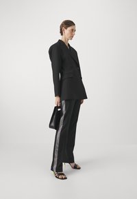 Black tailored blazer with structured shoulders, paired with black pants featuring a side rhinestone stripe. Carrying a small black bag.