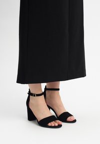 Crne suede sandale na petu s remenom oko gležnja i zlatnom kopčom, otvoreni dizajn prstiju, s teksturiranom površinom i debelom petom.