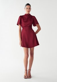 Bordo satino suknelė su aukšta apykakle ir trumpomis pūstomis rankovėmis, turinti prigludusią juosmenį ir išskirtą sijoną. Juodos dirželinės kulniukės užbaigia šį įvaizdį.