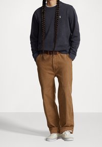 Mörkgrå långärmad sweatshirt, bruna tapered byxor med sidofickor och ljusa sneakers. Sweatshirten har en liten broderad logga.