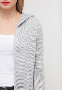 Světlé šedé pletené sako s kapucí, s volným pletením a dlouhými rukávy, nošené přes bílý tílko. Hladká textura, ležérní design.