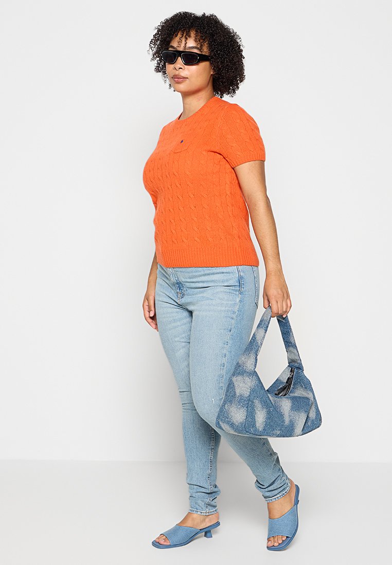 Maglione corto arancione a maniche corte con texture a treccia, jeans azzurri, sandali con tacco blu e una borsa a tracolla blu con fantasia.